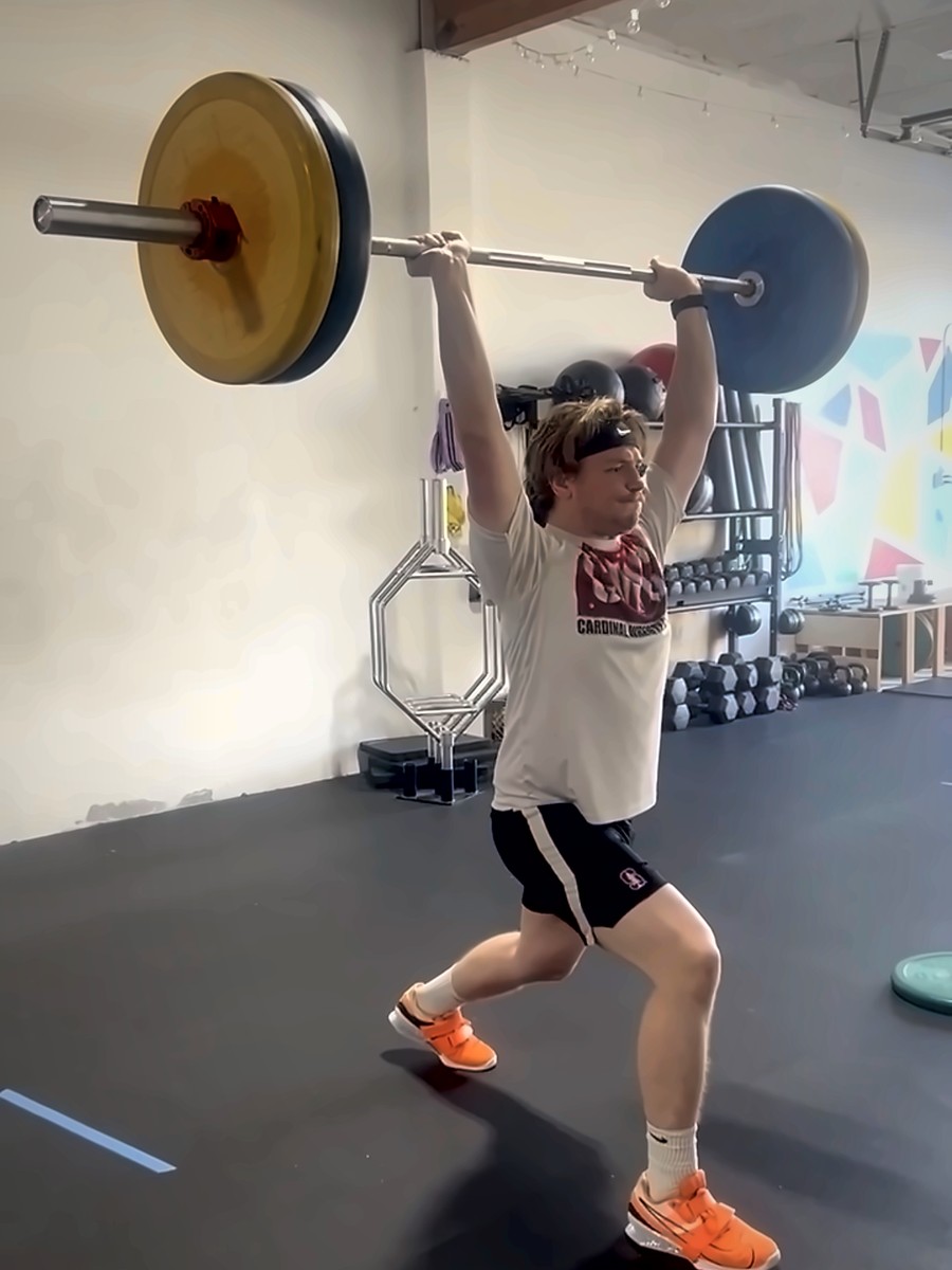 An adult athlete locking out a barbell overhead in the split position at Virtus